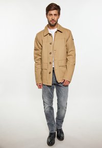 Beige bomuldsjakke med krave, knaplukning og to forlommer, parret med grå jeans og sorte sko.