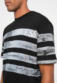 Čierne tričko s krátkym rukávom, s horizontálnymi sivými pruhmi a bielym logom "GUESS", s jemnou textúrou a štandardným strihom.