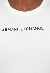 Bela bombažna majica s kratkimi rokavi z izvezenim logotipom "Armani Exchange" v črni barvi. Klasična zasnova s krožnim izrezom in gladko teksturo.