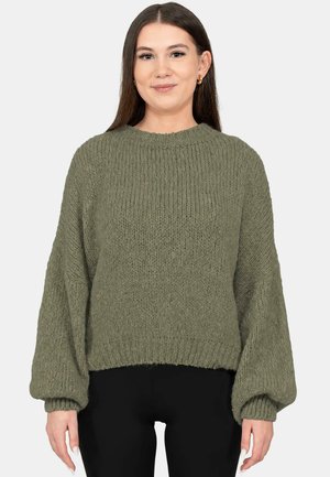 Femme portant un pull en maille vert olive ample avec des manches ballon et un pantalon noir, debout devant un fond uni.