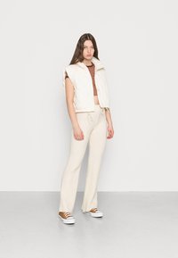 Colete acolchoado branco, curto, com textura de quilt, top curto às riscas, calças de malha ribana beje suave e ténis castanhos com solas brancas, a estar em pé num chão cinza.