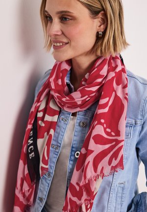Glimlachende vrouw met kort blond haar, die hoepeloorbellen, een licht denimjack en een roze-rood sjaal met patroon om haar hals draagt.