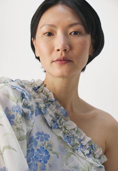 Eén-schouder blouse van lichtgewicht, doorzichtig materiaal met bloemmotieven in blauw en groen, benadrukt door geraffineerde details langs de rand.