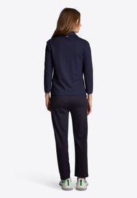 Kék zipzáras dzseki, gallérral, hosszú ujjakkal és gomb részlettel a hátsó részen, párosítva hozzáillő slim-fit nadrággal és sportcipővel.
