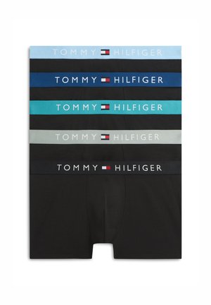 Jeune homme debout pieds nus portant un boxer noir Tommy Hilfiger, souriant avec les mains jointes devant lui sur un fond blanc.