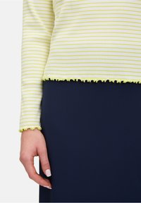 Gelb- und weiß gestreifte Bluse mit langen Ärmeln, die einen fließenden Saum und Manschetten aufweist. Kombiniert mit einer einfarbigen navyblauen Hose.