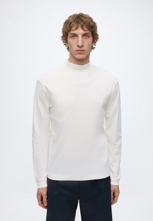 Junger Mann mit lockigem hellbraunem Haar, der ein schlichtes weißes Langarmshirt und dunkle Hosen trägt, steht vor einem weißen Hintergrund.