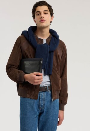 Ung mand iført brun ruskindjakke, blå tørklæde draperet over skuldrene, holder sort læderpung, iført lysegrå skjorte og blå jeans.