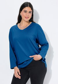 Niebieski, długi rękaw, top z dekoltem w serek wykonany z miękkiej tkaniny, z luźnym krojem i rozcięciami po bokach, zestawiony z ciemnymi obcisłymi dżinsami. Codzienny look.