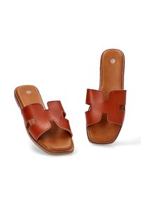 Sandalias de cuero marrón tipo zueco con la punta abierta y un diseño de tira cruzada. Presentan una textura suave y detalles de costura a lo largo de los bordes.