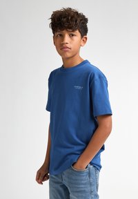 Camiseta de algodón azul de manga corta, con un pequeño logo blanco en el frente. Combinada con vaqueros azules desgastados.