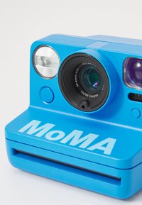 Blue instant camera with a prominent lens, built-in flash, and large letters spelling "MoMA" on the front. Smooth, plastic texture.