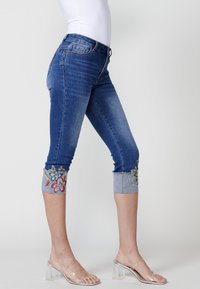 Jean capri en denim bleu foncé avec broderies florales aux poignets bleu clair. Taille ajustée avec une décoloration et un ourlet brut. Comprend une poche avant.