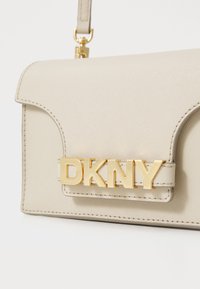 Bežinė krepšys su tekstūruotu paviršiumi, su ryškiai matomu auksiniu "DKNY" logotipu ir užsegimu su voleliniu siuvinėjimu.