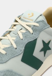 Light blue sneaker featuring textured grey suede accents, a green leather logo, and cream laces. The tongue has a gold "Converse" logo.