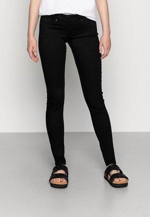 Persoon die zwarte skinny jeans en zwarte sandalen draagt, staand op een grijze vloer tegen een witte muur.