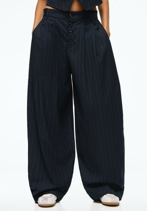 Pantalon noir ample à fines rayures, taille haute avec des boutons sur le devant, porté avec des chaussures blanches et une veste courte assortie.