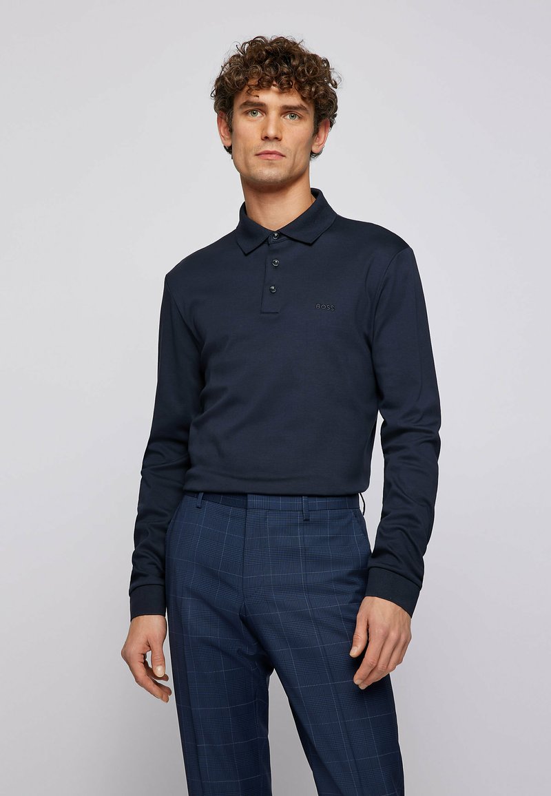 Navy long-sleeve polo shirt with a collar, three buttons, and a subtle logo. Paired with checked trousers in darker blue tones.