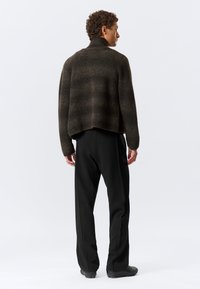 Rjav pleteni pulover z visokim ovratnikom in širokim krojem, v kombinaciji s črnimi hlačami. Pletež ima teksturirani vzorec in skrajšan zadnji del.