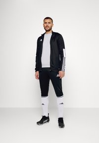 Zwart atletisch trainingspak met witte strepen, aansluitend design. Gedragen met een grijs shirt, zwarte voetbalschoenen en witte sokken met zwarte accenten.