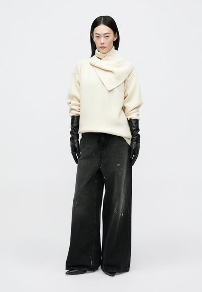 Krem pulover z ribasto teksturo in širokim zavihkom, v kombinaciji z širokimi črnimi kavbojkami z barvnimi madeži, dopolnjenimi s črnimi usnjenimi rokavicami.