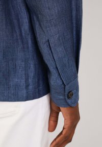 Manica di camicia con motivo a spina di pesce blu e chiusura a bottone al polsino, realizzata in un tessuto testurizzato, abbinata a pantaloni bianchi.