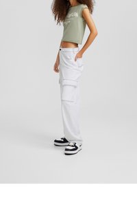 Camiseta verde recortada con texto blanco, pantalones cargo blancos con bolsillos y zapatillas en blanco y negro. Tejido suave, corte relajado.