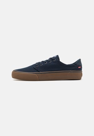 Zapatillas de lona azul marino con punta redonda, diseño de cordones, suela de goma en color marrón y etiqueta de logo en el lateral. Textura suave con un detallado mínimo.