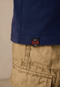 Tumši zila kokvilnas krekla ar sarkanu un melnu "Superdry" birku, apvienota ar bežām cargo šortiem, kuros ir izšūti detalējumi un kabatas.