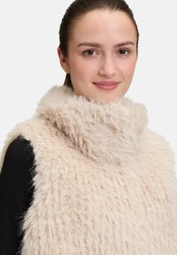 Gilet en fausse fourrure beige avec un col haut, présentant une surface douce et texturée ainsi qu'une coupe légèrement cintrée, porté par-dessus un haut noir.