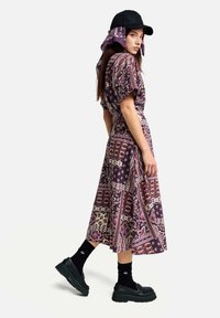 �Žena v midi šatech fialové barvy s květinovým patchworkovým vzorem, černé čepici s odpovídajícím šátkem, černých ponožkách a robustních černých botách na platformě.