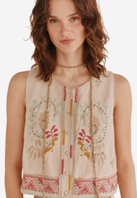 Top sans manches beige brodé avec des motifs floraux en rose, jaune et bleu, accompagné de boutons et d'accents en perles le long de l'ourlet.