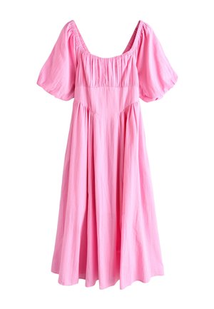 Ung kvinde i pink kjole smiler med korslagte arme, stående ved siden af et hvidt vintage terrængående køretøj på en bygade.