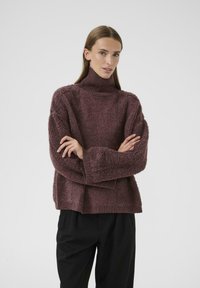 Frau mit langen Haaren, die einen strukturierten lila Rollkragenpullover und schwarze Hosen trägt, steht mit verschränkten Armen vor einem schlichten Hintergrund.