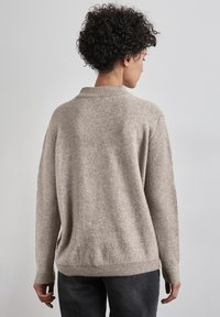 Maglione beige lavorato a maglia con una texture a coste, caratterizzato da un collo alto, spalle scese e polsini a coste. Indossato sopra jeans scuri.