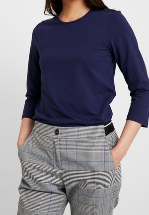 Femme portant un haut bleu marine à manches longues avec des boutons dorés sur les épaules et un pantalon taille haute à carreaux gris, les mains dans les poches.