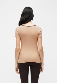 Femme aux longs cheveux noirs portant un haut beige à manches courtes et un pantalon noir, vue de dos sur un fond uni.