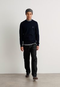 Navy knitted sweater, gray collared shirt underneath, dark jeans, and gray beanie. Model stands against a light backdrop, showing casual outfit details.