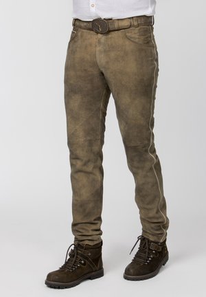 Braune Wildlederhose mit weißem Seitenstreifen, kombiniert mit braunen Schnürstiefeln und einem Gürtel mit Hirsch-Emblem-Schließe.