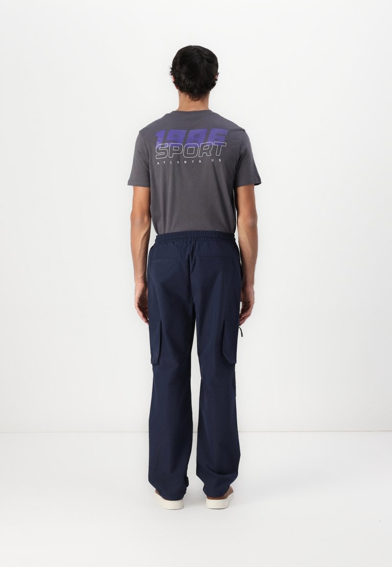 Homme portant un t-shirt gris avec le texte "1986 SPORT ATLANTA US" et un pantalon cargo bleu marine, debout face à un mur blanc.
