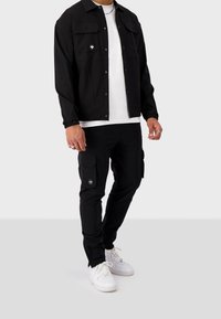 Czarna kurtka zapinana na guziki z dwoma kieszeniami na piersi, w połączeniu z czarnymi spodniami cargo z bocznymi kieszeniami oraz białymi trampkami.