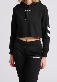 Hoodie nera cropped con cappuccio e coulisse, caratterizzata da testo bianco "HUMMEL". Abbinata a pantaloni della tuta neri coordinati, entrambi realizzati in morbido tessuto.