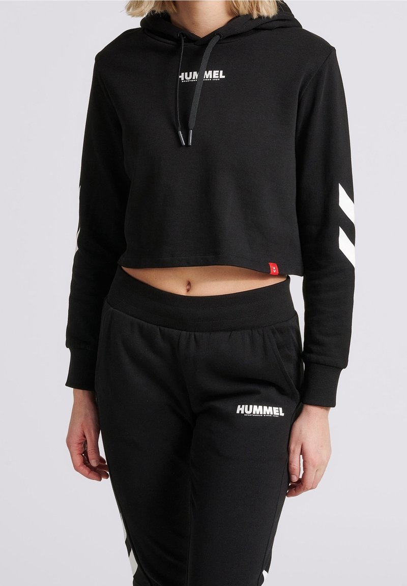 Hoodie nera cropped con cappuccio e coulisse, caratterizzata da testo bianco "HUMMEL". Abbinata a pantaloni della tuta neri coordinati, entrambi realizzati in morbido tessuto.