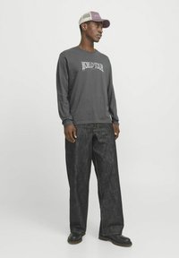 Graues Langarmshirt mit "WORLD TOUR"-Grafik, kombiniert mit locker sitzenden dunklen Jeans und schwarzen Stiefeln, vor einem neutralen Hintergrund.