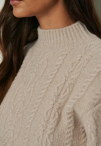 Pull beige tricoté avec un col montant côtelé, présentant des motifs torsadés et une finition douce et texturée, vu de profil.
