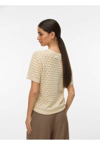 Femme aux cheveux longs et foncés attachés en queue de cheval basse, portant un haut en maille crème à manches courtes avec un motif en forme de cœur et un pantalon marron, vue de dos.