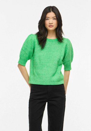 Femme aux longs cheveux foncés portant un pull vert vif à manches bouffantes et un pantalon noir, debout les mains derrière le dos.