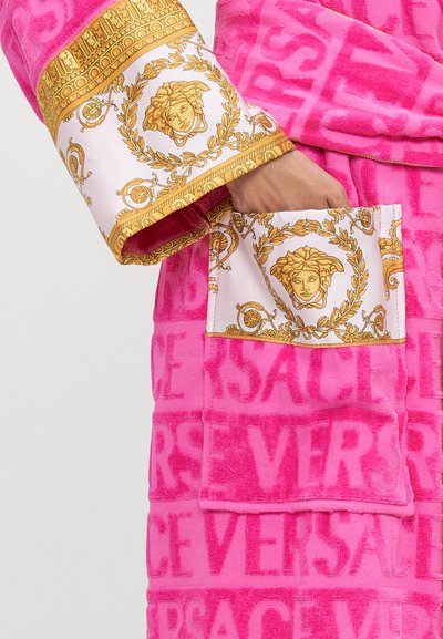 Rozā frotē halāts ar zelta akcentiem pie piedurknēm un kabatu, rotāts ar Versace motīvu, kas ietver dekoratīvu medaļu.