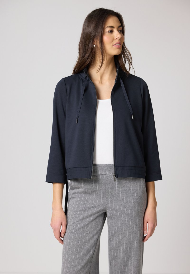 Donna che indossa una giacca con zip blu navy con coulisse, top bianco e pantaloni grigi a righe sottili, che guarda verso destra su uno sfondo semplice.