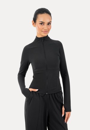 Mujer con cabello oscuro recogido en una cola de caballo elegante, vistiendo una chaqueta negra ajustada con cremallera y pantalones negros sueltos, posando con una mano en el bolsillo.